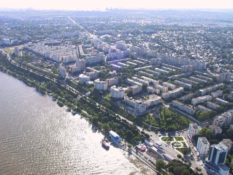 An aerial daytime view of the city of Galați, Romania, situated next to a wide, sunlit river. The cityscape features a dense mix of apartment blocks, commercial buildings, and green, tree-lined areas stretching inland from the waterfront toward a hazy horizon.