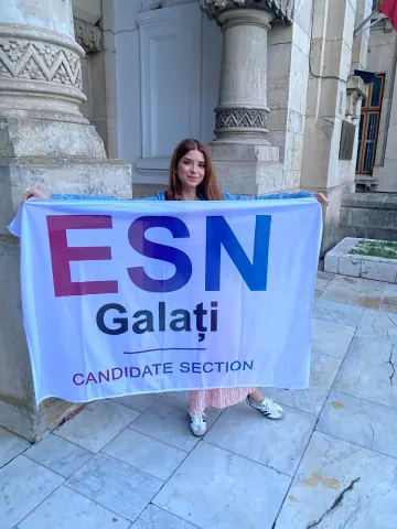 A young woman standing outdoors on a stone-paved area, holding up a large white flag. The flag displays 'ESN' in large pink and blue letters, with 'Galați' printed below it in dark blue, and the words 'CANDIDATE SECTION' in smaller text at the bottom. She is positioned in front of a building with classic stone pillars.