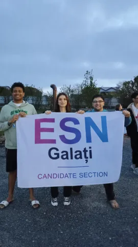 Three young adults standing outdoors on a paved area, jointly holding up a large white flag. The flag displays the letters 'ESN' in large pink and blue text, with 'Galați' printed below it in dark blue, and the words 'CANDIDATE SECTION' in smaller text at the bottom.