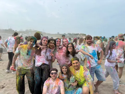 A lively group photo of several young adults outdoors on a sandy beach, all smiling and covered head-to-toe in brightly colored powder, typical of a Holi festival or color run. The main group is clustered together in the foreground, with three people kneeling and several standing behind them, their clothes stained with pink, yellow, blue, and green powder. Other participants can be seen in the background enjoying the beach under a light, overcast sky.