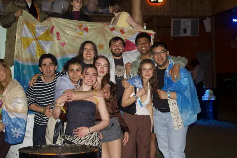 A group photo of approximately nine young adults smiling and posing together at an dimly lit indoor party or social gathering. They are dressed casually and standing closely, with their arms around each other's shoulders. Draped over a structure in the background behind them is a large flag featuring white, blue, and yellow vertical stripes.
