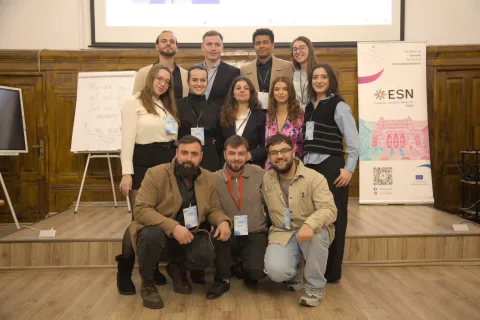 A group photo of 12 people posing together on a small indoor wooden stage. The group is arranged in three rows, with the front three individuals crouching and the rest standing behind them. Many in the group are wearing large event badges on lanyards. The room features wood-paneled walls, with a projection screen visible at the top, a whiteboard easel to the left, and a vertical ESN event banner standing to the right.