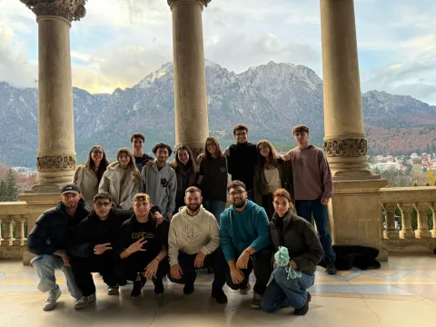 A group photo of approximately 15 to 20 young adults posing together on a covered stone terrace. The group is casually dressed in cool-weather clothing and arranged in two rows, with the front row crouching and the back row standing. They are framed by large, classical stone columns, with a scenic view of a prominent mountain range in the background beneath a partly cloudy sky.